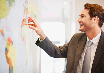 Imagem 482x300 Outros Negócios Fotografia de um homem sorrindo e apontando para um mapa mundo na parede.