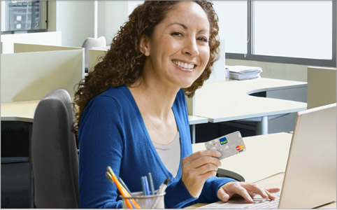Fotografia de uma mulher sorridente segurando um cartão de crédito do Itaú na cor cinza sentada em frente à uma escrivaninha com seu computador em cima.