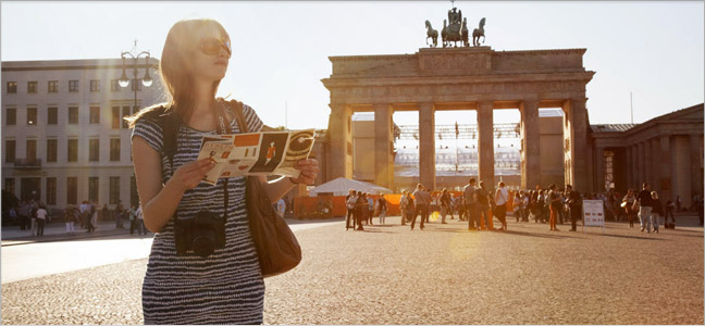 Viagens ao exterior Imagem de uma mulher passeando num ponto turístico com um mapa em suas mãos e uma câmera pendurada em seu pescoço. Aproveite suas férias com viagens ao exterior.