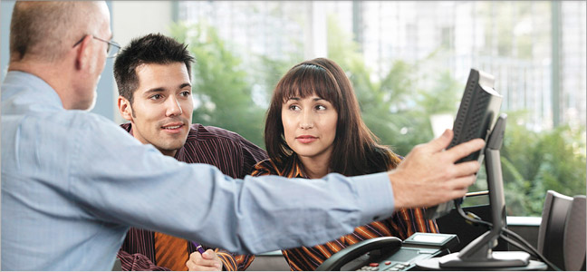 banner Fotografia de um casal conversando com um gerente que aponta para o computador a sua frente.