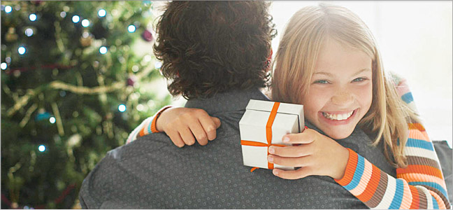 imagem 648x300 Fotografia de menina abraçando o seu pai enquanto segura uma caixinha de presente branca com um laço na cor laranja.