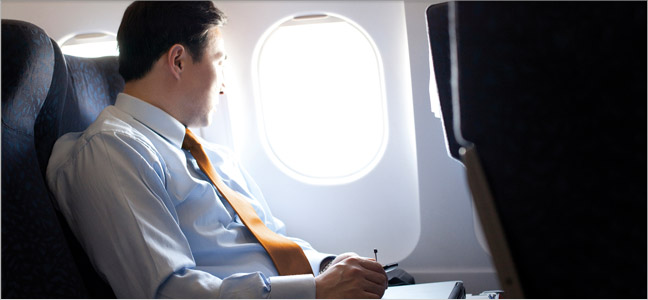 Imagem - Seguro viagem Fotografia de um homem vestido de camisa e gravata dentro de um avião olhando pela janela.